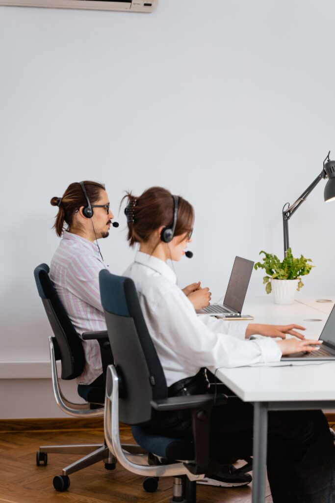 Contact Us Two call center employees working with laptops and headsets in an office.